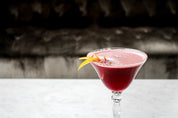 Red cocktail with a lemon twist in a martini glass on a white surface.