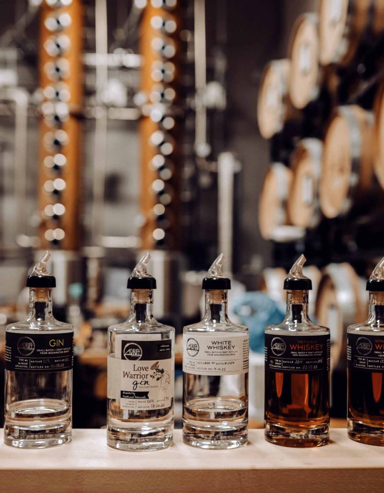 Five bottles of gin and whiskey on a wooden surface with a blurred background of barrels.